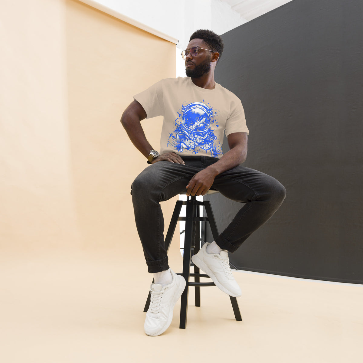 Model seated on a stool wearing a sand Blue Astronaut graphic tee, dark trousers and white high-top shoes